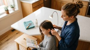 An Omaha-area parent gently lifts a section of her elementary-school-aged daughter's freshly-combed brown hair near the nape of the neck to inspect the scalp at the kitchen counter, with a fine metal nit comb, a folded teal hand towel, and a bottle of conditioner ready beside them.
