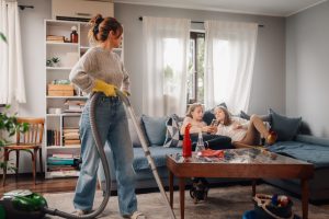 Parent vacuuming a fabric living room couch with two daughters sitting on the sofa during a household cleanup