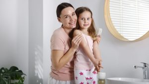 Mother brushing her young daughter's freshly washed hair in a softly lit bathroom, the canonical decision moment when a parent looking at a freshly clean head wonders whether hair hygiene affects whether head lice will infest it.