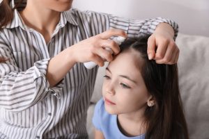 Mother holding an aerosol styling spray near her young daughter's hair in a softly lit bathroom, the canonical decision moment when a parent considers reaching for a household styling product to deal with head lice.