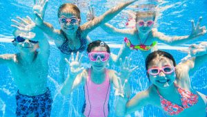 Children swimming underwater in a sunny outdoor pool - the decision-moment scene parents picture when wondering whether pool chlorine kills head lice or whether their kids can catch lice at the pool this summer.
