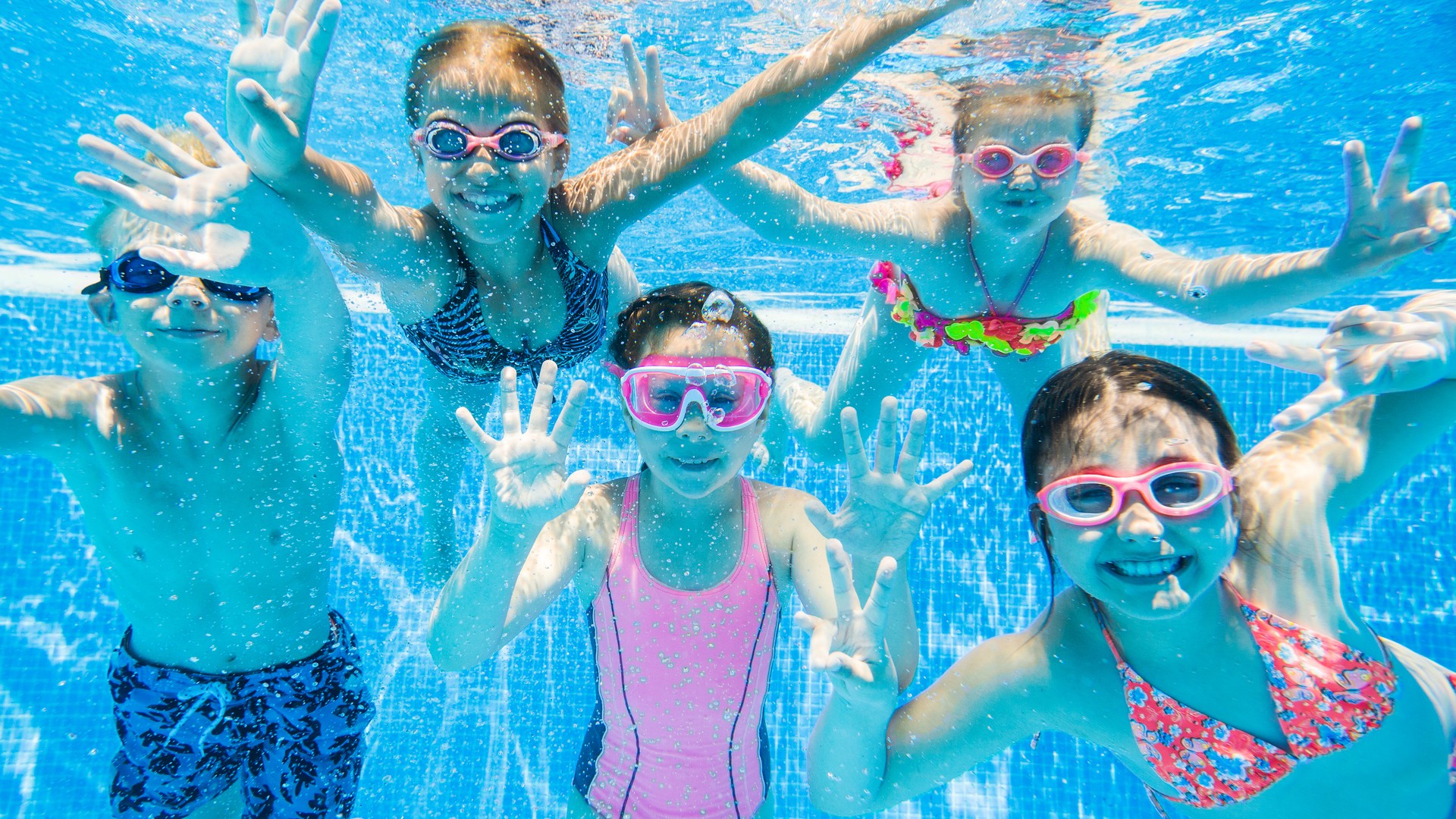 Children swimming underwater in a sunny outdoor pool - the decision-moment scene parents picture when wondering whether pool chlorine kills head lice or whether their kids can catch lice at the pool this summer.