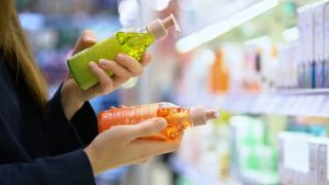 Close-up of a mother's hands comparing two hair-care bottles in a cosmetics-department aisle, the exact drugstore-shelf moment an Omaha parent faces when deciding between a lice-treatment mousse and the shampoo sitting next to it.