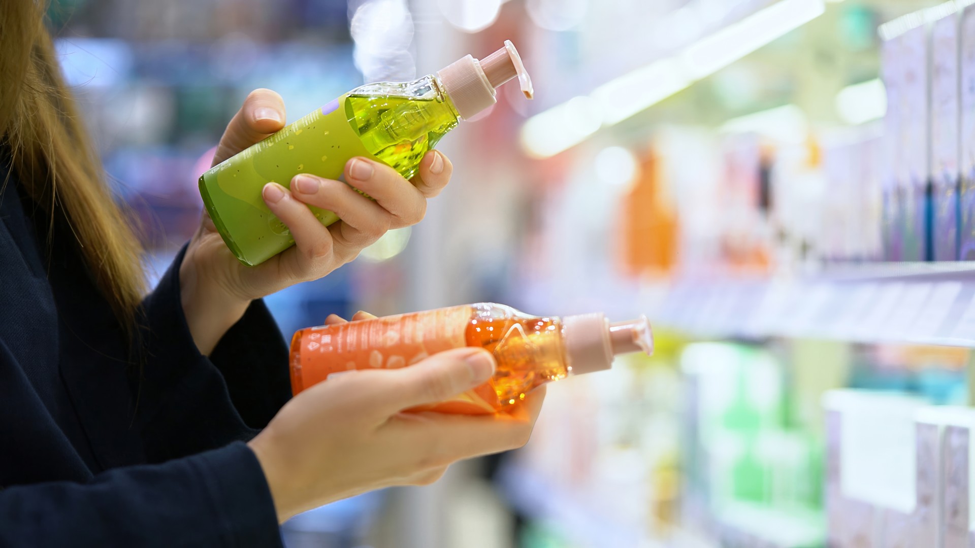 Close-up of a mother's hands comparing two hair-care bottles in a cosmetics-department aisle, the exact drugstore-shelf moment an Omaha parent faces when deciding between a lice-treatment mousse and the shampoo sitting next to it.