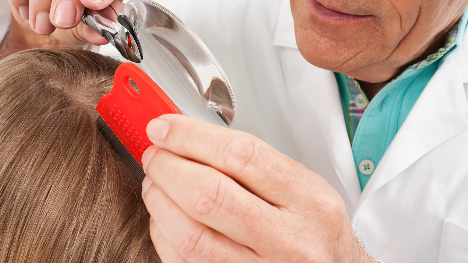 Clinician combing a child's hair with a red fine-toothed lice comb while inspecting it under a magnifier