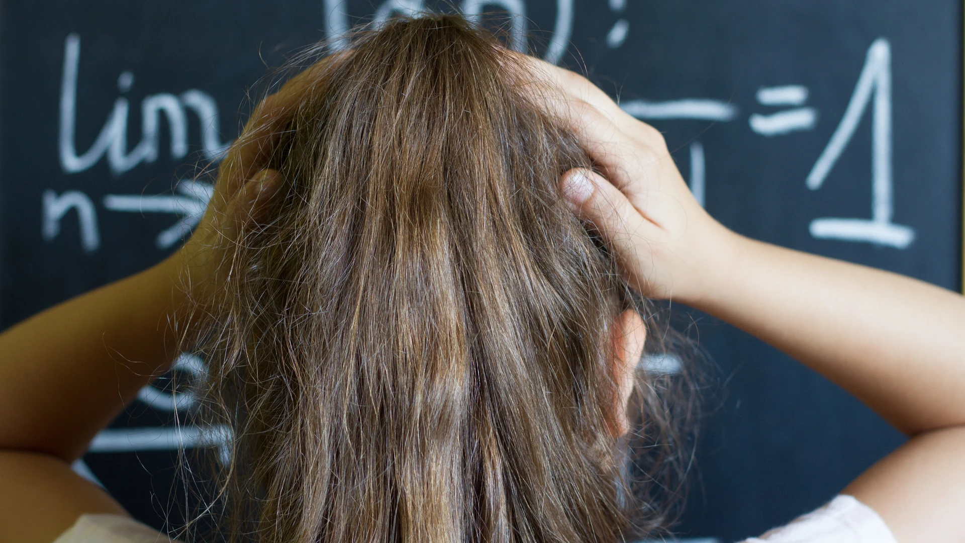 A girl touches the back of her head in front of a classroom chalkboard.