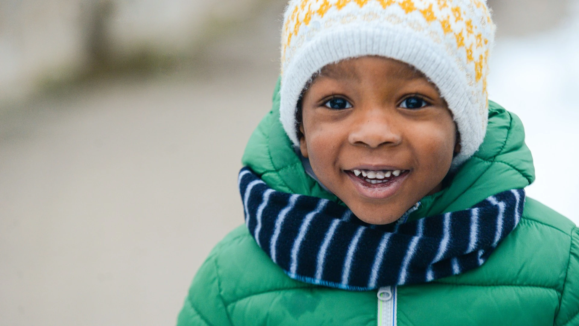 A smiling child wears a knit winter hat and scarf outdoors.