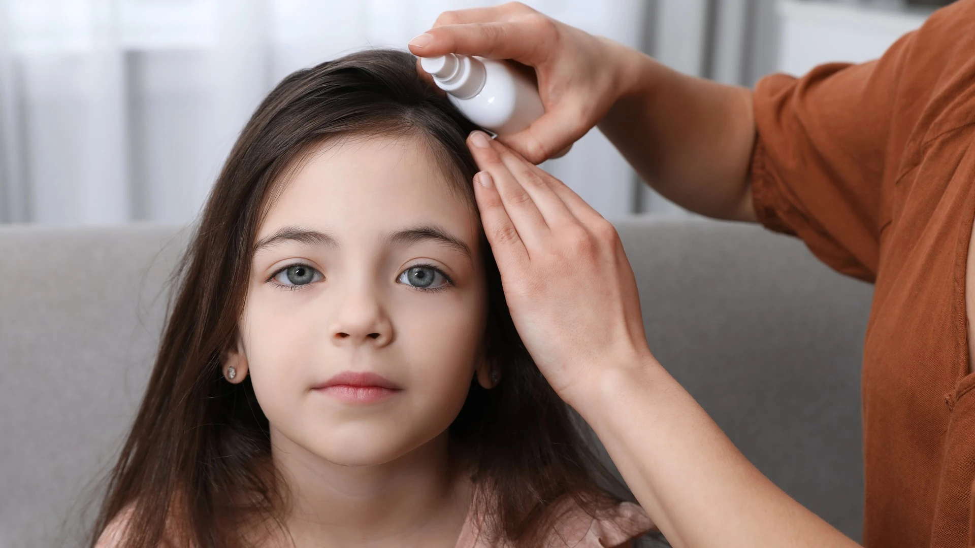 A mother sprays her daughter's hair near the hairline indoors.