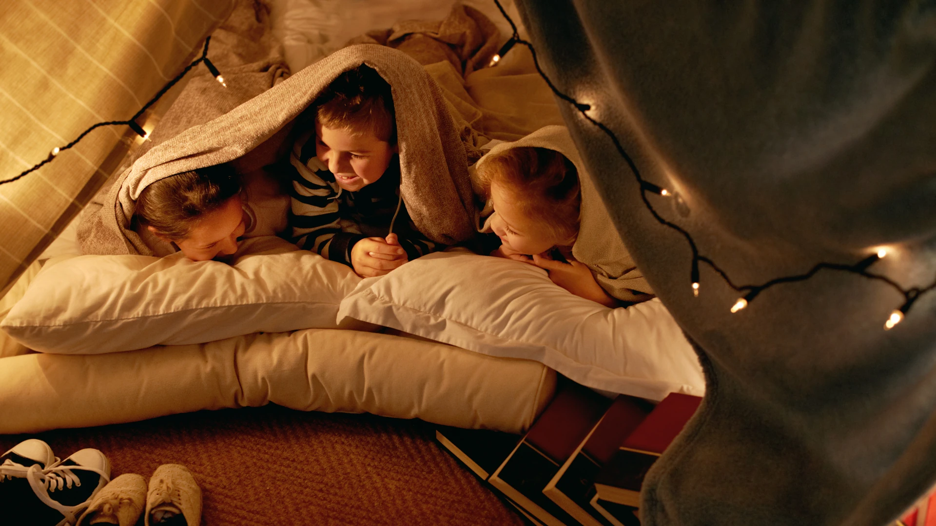 Children rest together inside a blanket fort strung with lights.