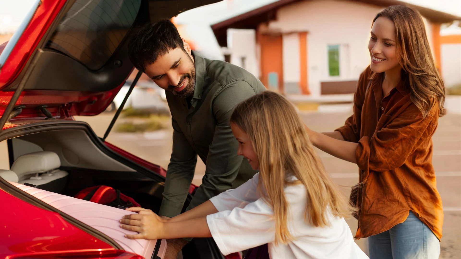 Two parents and their daughter load luggage into a car trunk before a trip.