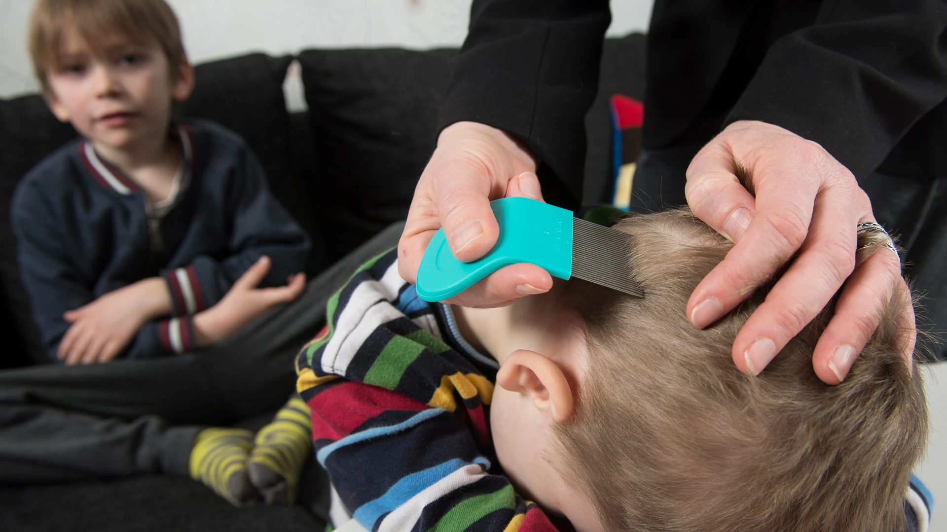 An adult combs one child's hair with a lice comb while another child waits nearby.